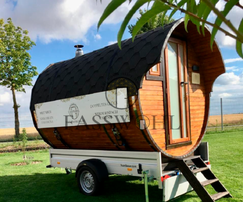 Eine Fasssauna steht auf grünem Rasen unter Bäumen