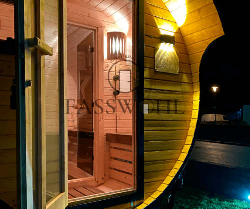Ein beleuchtetes Fasswohl Saunafass steht mit offener Tür im Garten