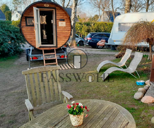 Im Vordergrund steht ein runder Tisch mit gemütlichen Stühlen aus Holz und im Hintergrund die Fasssauna