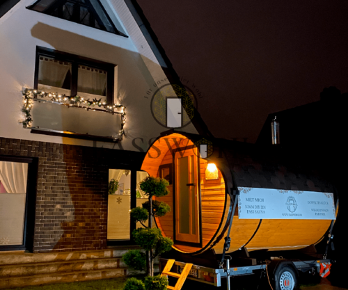 Eine weihnachtlich beleuchtete Fasssauna bei Nacht im Garten vor einem beleuchteten Haus